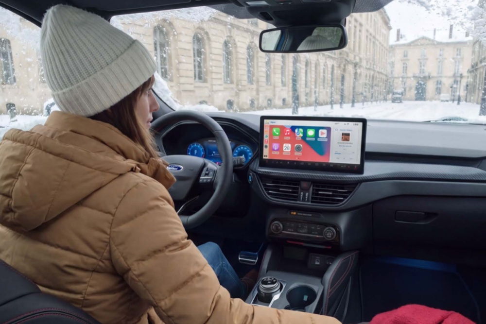 Eine Frau in Wintermantel und Haube sitzt an einem verschneiten Tag in einem Ford und greift nach einer roten Decke.