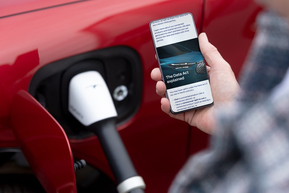 Eine Person checkt ihr Smartphone, ein rotes Elektrofahrzeug lädt im Hintergrund auf.