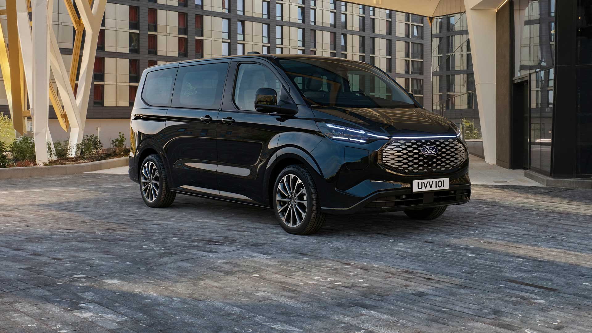 Ford E-Tourneo Custom in Schwarz. Dreiviertelansicht, parkt vor einem modernen Gebäude.