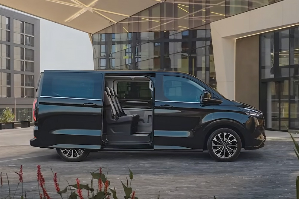 Ford E-Tourneo Custom in Schwarz. Seitenansicht, parkend vor modernem Gebäude. 