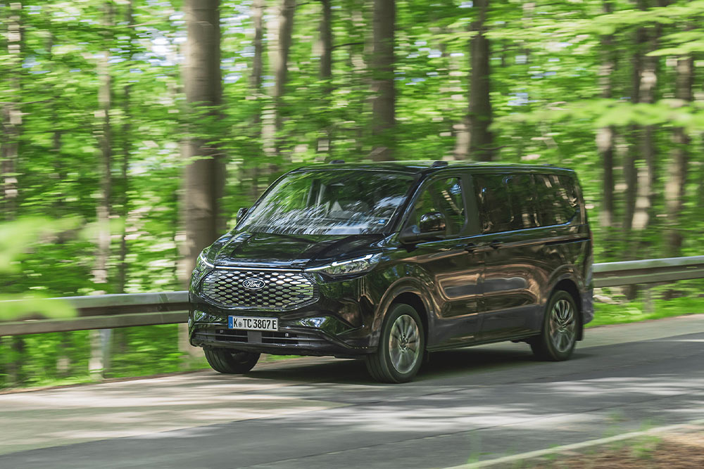 Ford E-Tourneo Custom in Schwarz. Dreiviertelansicht, fährt auf einer Straße in einem bewaldeten Gebiet.