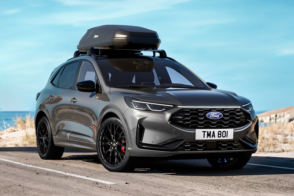 Vorderansicht eines dunkelgrauen Ford Kuga mit Dachbox und schwarzen Rädern, am Meer geparkt.