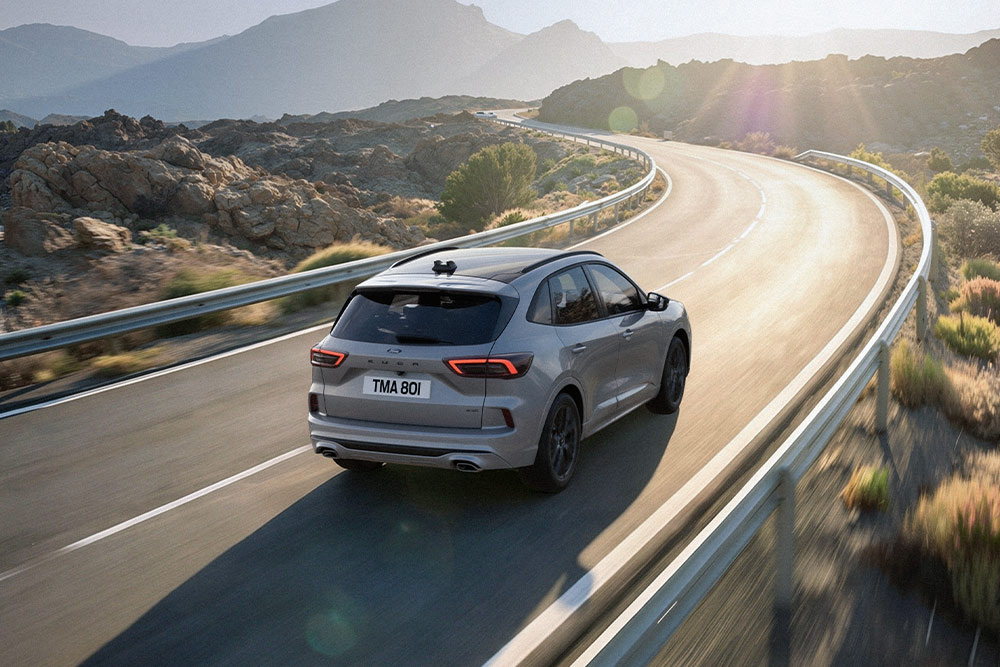 Ford Kuga in Silber. Heckansicht, bei der Fahrt auf einer kurvigen Straße durch eine sonnige Berglandschaft.