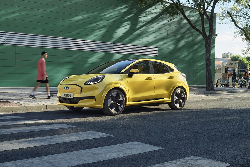 Ford Puma Gen-E in Gelb. Dreiviertelansicht, bleibt vor einem Zebrastreifen stehen, rechts davon ein grünes Gebäude.