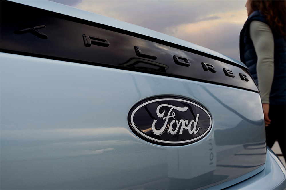 Ford Explorer in Blau. Nahansicht mit Zoom auf das Ford Logo. Person geht im Hintergrund am Fahrzeug vorbei. 