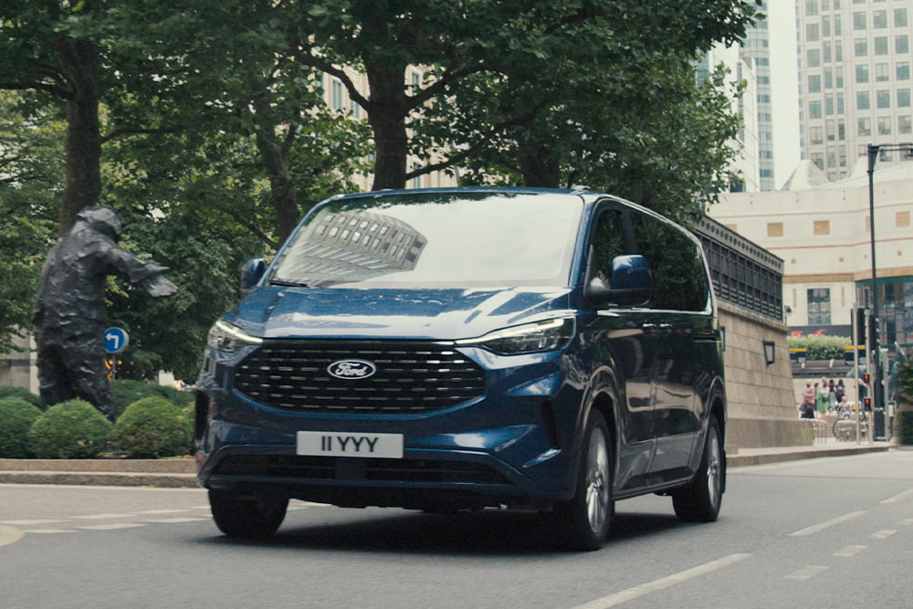 Ford Tourneo Custom in Blau. Dreiviertelansicht, fahrend auf einer Straße mit Hochhäusern und Bäumen im Hintergrund.