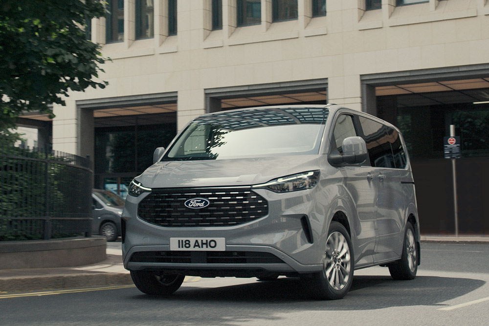 Ford Tourneo Custom in Grau. Dreiviertelansicht, fährt um die Kurve auf einer Straße mit weißen Gebäuden.
