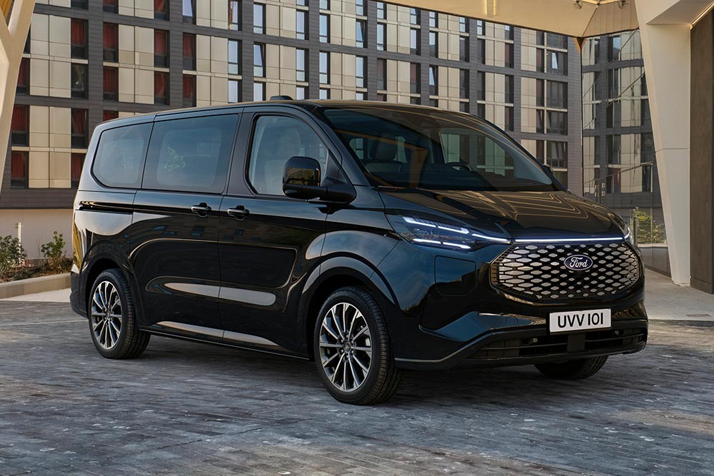 Ford E-Tourneo Custom in Schwarz. Dreiviertelansicht, parkt vor einem modernen Gebäude.