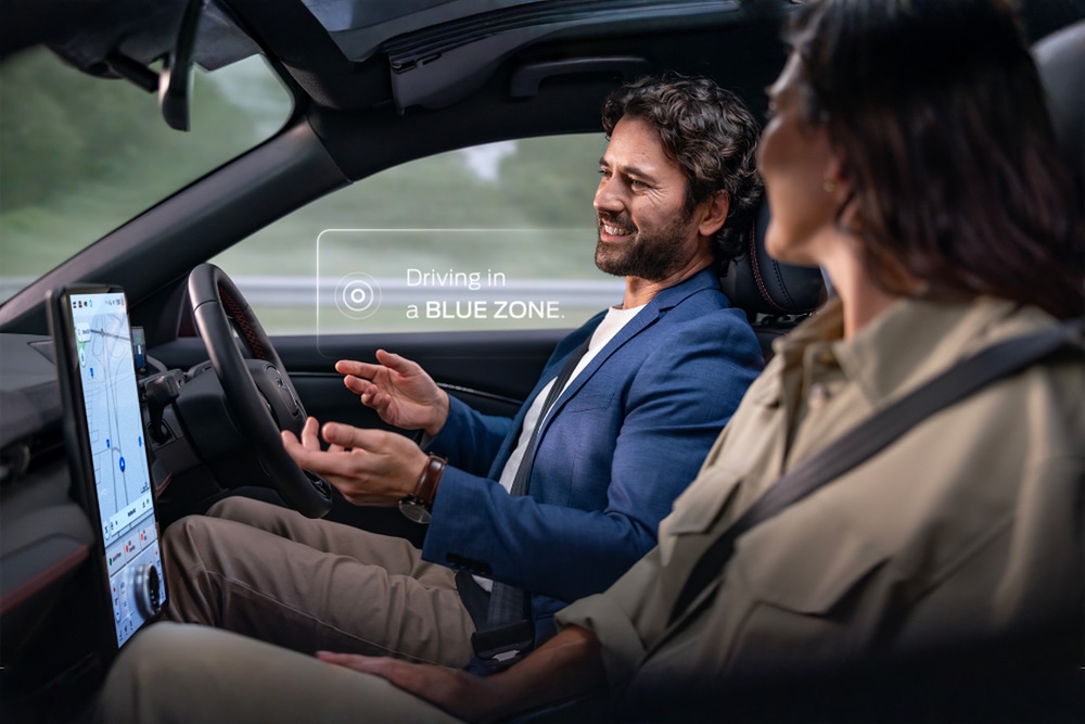 Zwei Personen sitzen im Ford Mustang Mach-E und fahren freihändig mit der Ford BlueCruise-Funktion. Ein Benachrichtigungsfester zeigt "Fahren in einer BLUE ZONE“ an.