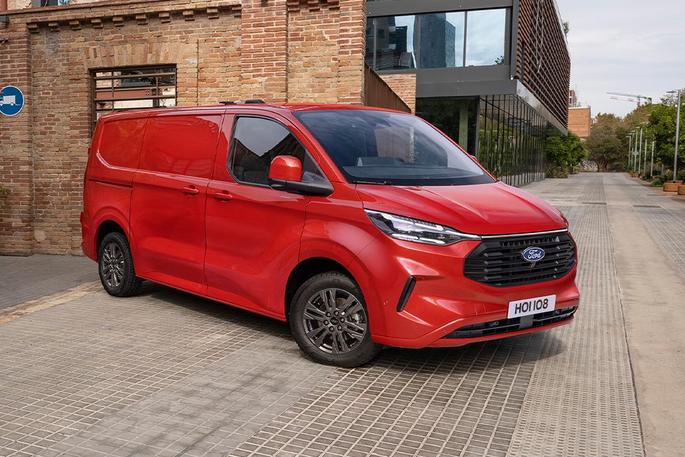 Ford Transit Custom in Rot. Dreiviertelansicht, parkt vor einem Ziegelsteingebäude.