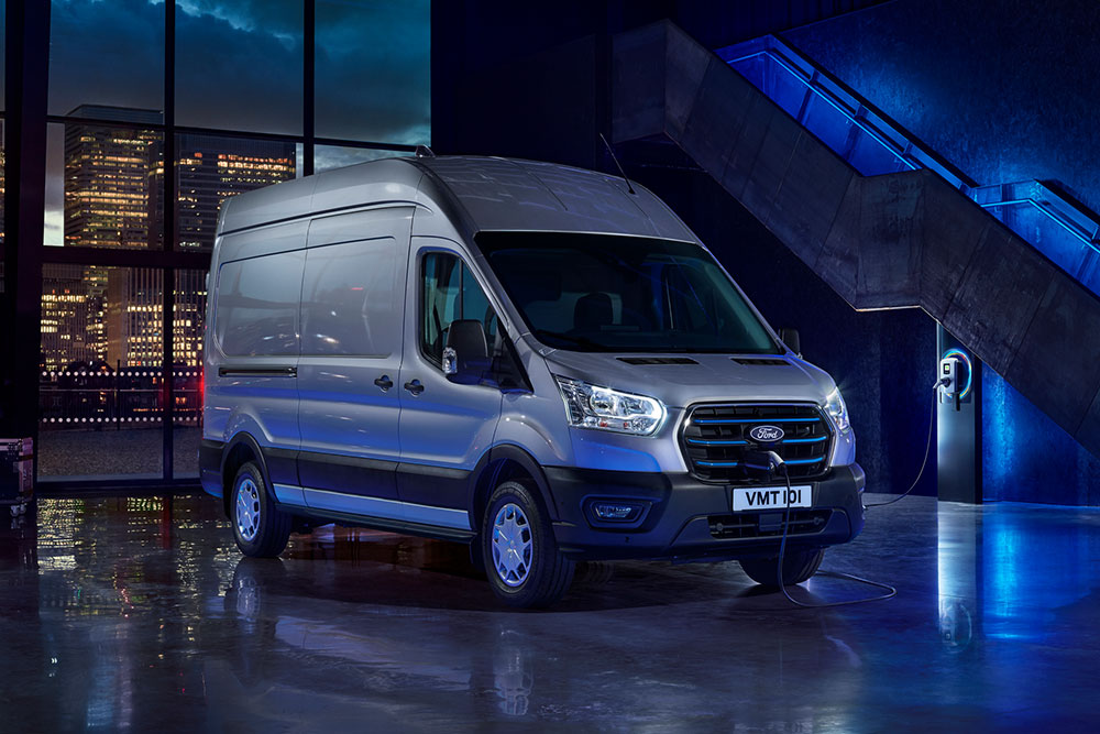 Ford E-Transit in Silber. Dreiviertelansicht, lädt an einer Wallbox in einer Lagerhalle.