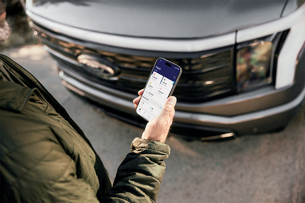 Hand hält ein Mobiltelefon mit geöffneter Ford App. Im Hintergrund ist ein Ford Transporter zu sehen.