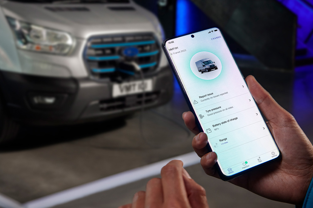 Person hält Smartphone mit geöffneter Ford App in der Hand, im unscharfen Hintergrund parkt ein silberner Ford Transit.
