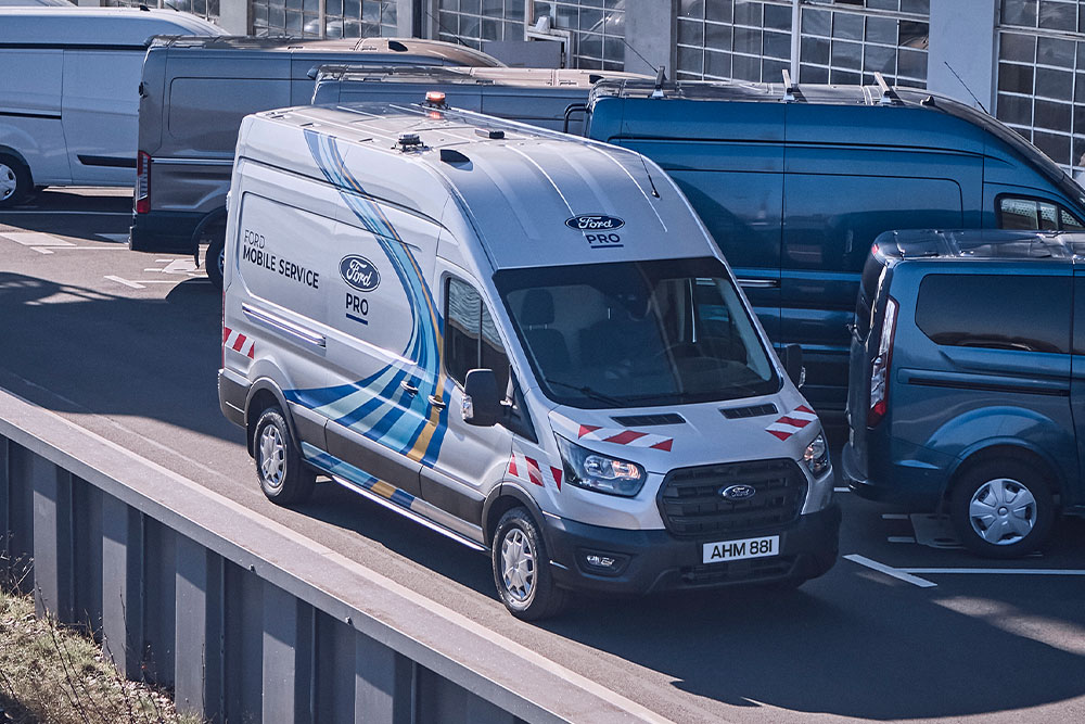 Ein silberner Ford Transit des Ford Pro Mobilen Service fährt auf einem Parkplatz. Im Hintergrund parken weitere Ford Fahrzeuge.