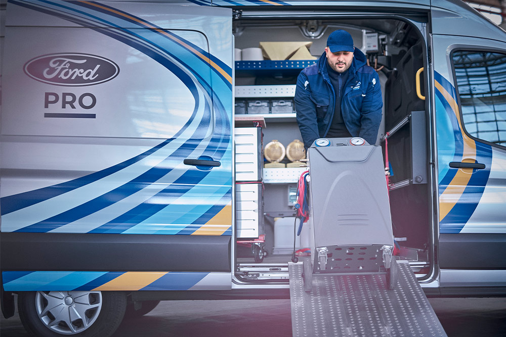 Ein Ford Mobiler Service Van, seitliche Ansicht, mit geöffneter Schiebetür. Ein Ford Pro Mitarbeiter transportiert Werkzeuge aus dem Transit-Transporter.