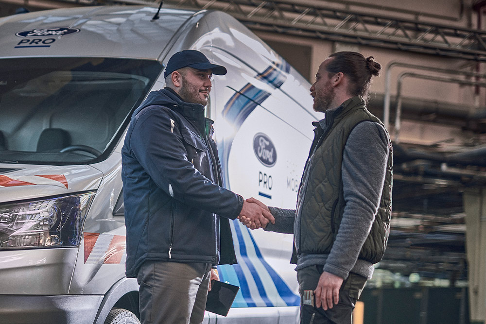 Ein mobiler Ford Pro-Techniker im Gespräch mit einem Kunden in einer Lagerhalle. Im Hintergrund ein Ford Pro Mobiler Service Transit Transporter.